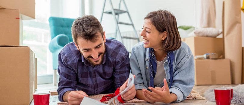 Couple in love moving in together, lying on the floor of their new home, searching for interior design ideas in a home decoration magazines and drinking coffee